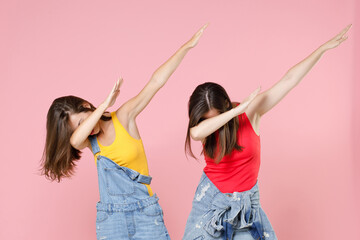 Wall Mural - Two cheerful funny young women friends 20s in casual denim clothes doing dab hip hop dance hands gesture, youth sign hiding and covering face isolated on pastel pink colour background studio portrait.