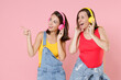 © ViDi Studio - Two funny young women friends 20s in casual denim clothes listening music with headphones pointing index finger aside on mock up copy space isolated on pastel pink colour background studio portrait.