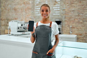 Young attractive woman barista stands at the counter in a coffee shop and smiles while showing a blank smartphone screen.