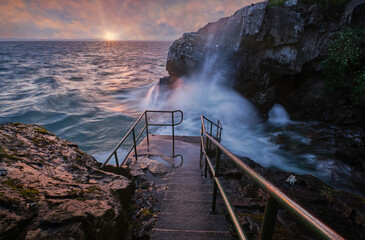 Wall Mural - Stairs lead down to the sea with big waves and sunset in the background