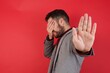 © Roquillo - Young caucasian businessman wearing casual clothes standing over isolated red background covers eyes with palm and doing stop gesture, tries to hide from everybody. People, body language.