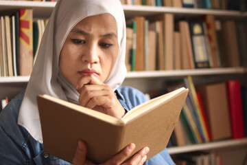 Wall Mural - Asian muslim woman wearing hijab reading book in library, educational concept. Happy smiling expression, doing leisure activity