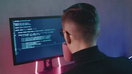 Rear view of man programmer coding network security software indoors in neon lights. Hacker typing code on computer screen in dark office at night