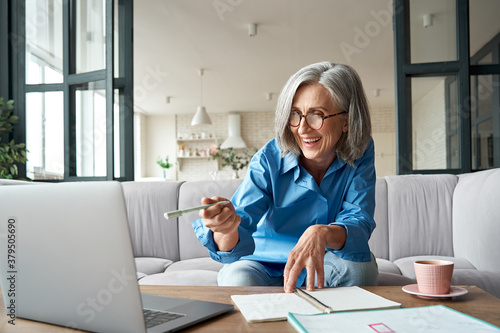 Fototapeta na szkło Happy mature older woman video calling on laptop working from home. Smiling 60s middle aged businesswoman talking by conference online virtual chat using computer at home office sitting on couch.
