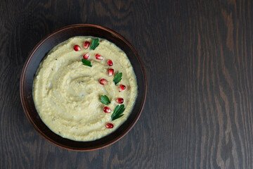 Wall Mural - Top view of single savory homemade vegetarian hummus made of chickpeas  decorated with pomegranate seeds and parsley served in brown bowl on wooden background at kitchen. Image with copy space