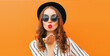 Portrait of stylish woman model blowing red lips sending sweet air kiss wearing a black round hat and striped white shirt on an orange background