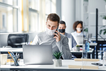 Social distance and online work with client remotely. Busy man in protective mask talking on smart phone sitting at office in workplace with laptop and glass board