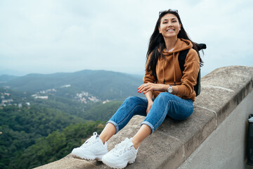 Smiling female traveler in casual wear sitting on destination outdoors satisfied with free time and journey, positive woman tourist looking at camera recreating on holiday vacation explore location