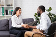 joyful patient shaking hands with african american psychologist while sitting on sofa