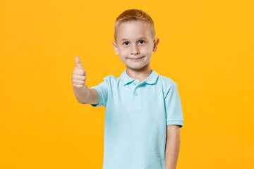 Wall Mural - Smiling little male fair-haired brown-eyed kid boy 5-6 years old wearing stylish blue turquoise t-shirt polo showing thumbs up gesture isolated on yellow color wall background, child studio portrait.