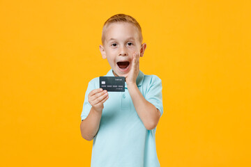 Wall Mural - Excited happy little fun male kid boy 5-6 years old wearing stylish blue turquoise t-shirt polo hold in hand mockups of credit bank card isolated on yellow color wall background child studio portrait.