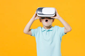 Wall Mural - Smiling handsome little fun male kid boy 5-6 years old wearing stylish blue turquoise t-shirt polo watching in vr headset gadget on head isolated on yellow color wall background child studio portrait.