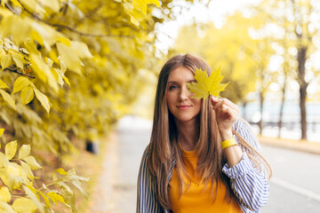 Portrait of happy cheerful woman with a smile holds an autumn yellow maple leaf near the face in the park on the nature walk outdoors