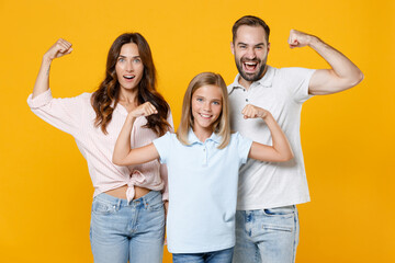 Wall Mural - Strong funny young parents mom dad with child kid daughter teen girl in basic t-shirts showing biceps muscles isolated on yellow background studio portrait. Family day parenthood childhood concept.