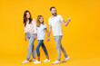 © ViDi Studio - Full length portrait of cheerful funny young parents mom dad with child kid daughter teen girl in t-shirts holding hands pointing index finger aside isolated on yellow background. Family day concept.