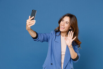 Wall Mural - Cheerful excited funny young brunette woman 20s wearing basic jacket doing selfie shot on mobile phone waving greeting with hand isolated on bright blue colour wall background, studio portrait.