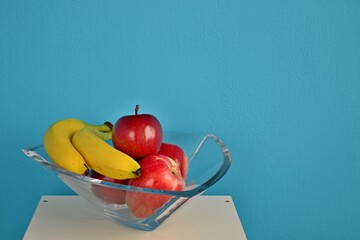 Fresh red apples and two bananas in a glass bowl. White table. Blue background. Still life with sweet fruit.