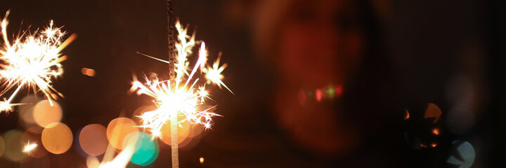 Wall Mural - Close-up of burning bengal lights. Smiling woman looking at sparkler with happiness and admiration on background. Happy New Year and Merry Christmas concept