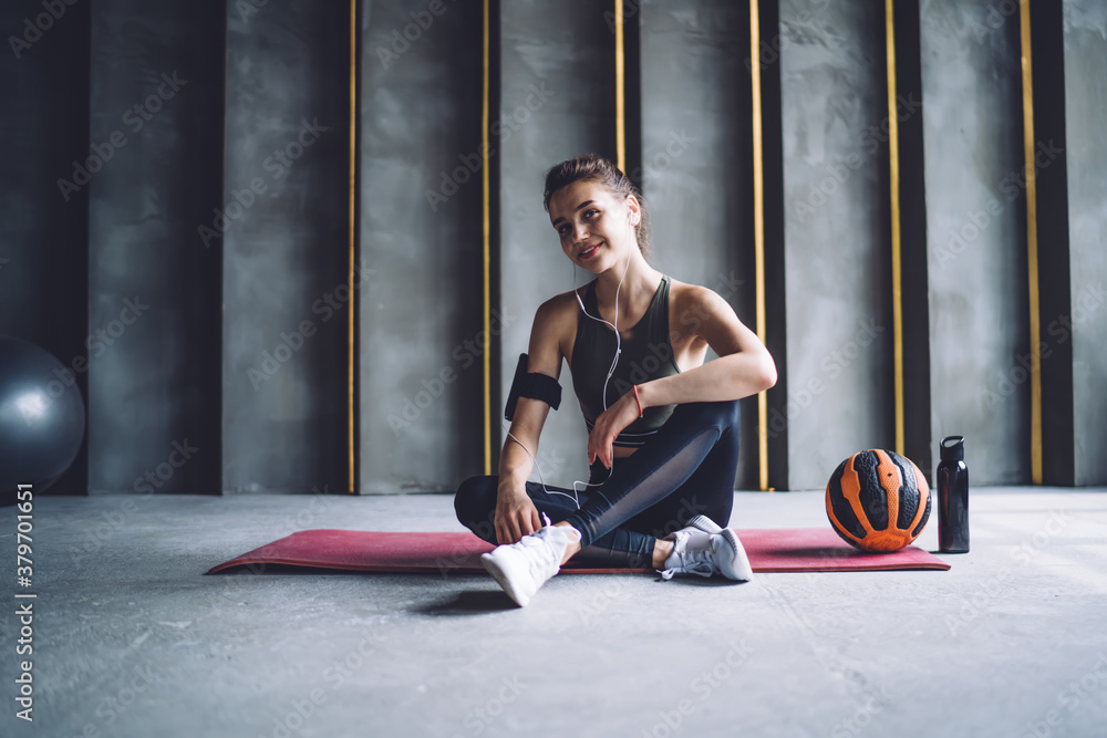 Half length portrait of professional prosperous woman in earphones resting after workout in gym, positive female athlete with ball looking at camera enjoying favorite songs from mobile phone - obrazy, fototapety, plakaty 