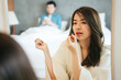 © THESHOTS.CO - Asian woman wearing bathrobe applying lipstick in bedroom.