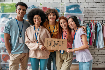 Canvas Print - Group of cheerful diverse young volunteers smiling at camera, holding card with Donate lettering while standing in charitable organization office
