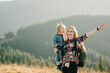 Mother with kid hiking in mountains. Young tourists on top of a mountain enjoying valley view before sunset. Happy family day. Mom with backpack hugging daughter.
