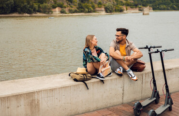 Wall Mural - Young couple with electric scooter on vacation having fun in the city, while sitting by the river.