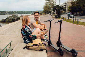 Wall Mural - Young couple with electric scooter on vacation having fun in the city, while sitting by the river.