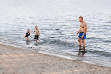 A Sports Man Of European Appearance Plays With His Children In The Water. The Family Bathes In The Lake. Father, Son And Daughter. Holidays And Games. Water Splash. Lakes In The Summer At Sunset.