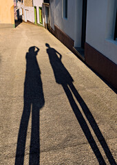 Canvas Print - Long shadows of two people in a narrow street of a village, one of them taking a picture, with nobody else in sight.