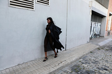 Sticker - Carefree ethnic young woman wears casual black coat, walking against urban background, focused into distance.