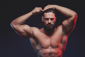 Muscular guy with black beard and mustache staying and combing his hairs in dark studio background.