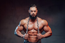 Brutal Athlete With Black Beard And Mustache And Naked Torso Holding Steel Chains And Posing With Them In Dark Background.