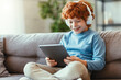 Delighted redhead boy with tablet sitting on sofa.