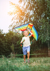 Wall Mural - Cheerful little boy running with a multicolored kite on the city park green grass meadow. Funny childhood concept image.