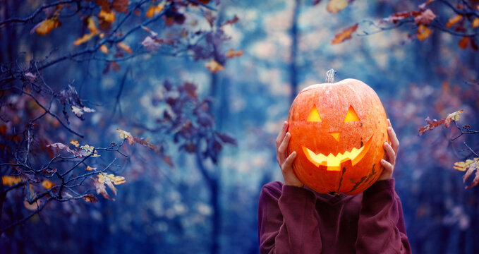 Boy holding carved pumpkin for halloween in front of his head in dark autumn forest. Halloween holiday concept.