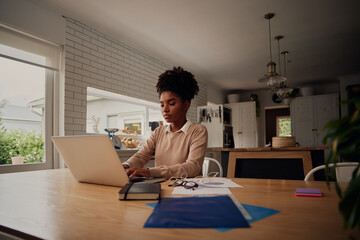 Wall Mural - Young concentrated african woman with curly hair working from home on computer while sitting at table at home