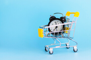 Alarm clock in mini shopping cart on blue background. Buying, time, sale concept. Copy space.