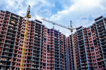 Very wide new brick apartment houses under construction. Two working cranes are nearby. It's possible to see stocks of construction materials on each floor.