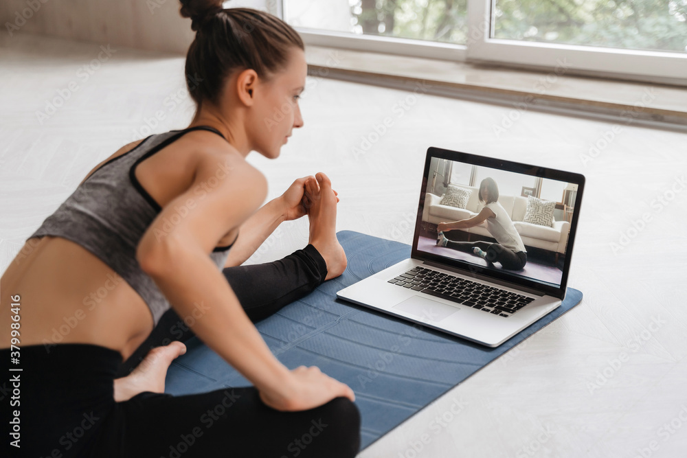 Fit woman in sportswear doing yoga on exercise mat at home using laptop watching tutorial. Female coach has a virtual yoga class on computer. Healthy and active lifestyle concept. Online home training - obrazy, fototapety, plakaty 