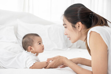 Beautiful asian mother and baby are playing on white bed and looking at each other. Happy asia mother and infant lying on bed at home.