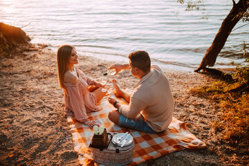 A beautiful blonde caucasian woman and a man are sitting on the beach drinking wine. A loving couple on a picnic by the river