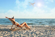 Young woman relaxing in deck chair on beach under sky with flying airplane. Summer vacation