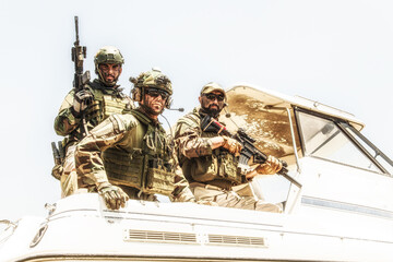 Army special operations soldiers, SEALs team, elite commando fighters group loaded with ammunition, wearing helmets and radio headset, armed assault rifles, standing together on speed boat stern