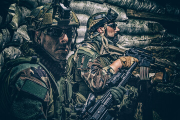 Soldiers covering from enemy fire in trench. Two Navy SEALs fighters, infantrymen in camouflage uniform and helmet, armed service rifles, hiding behind sandbags during night attack or gunfight