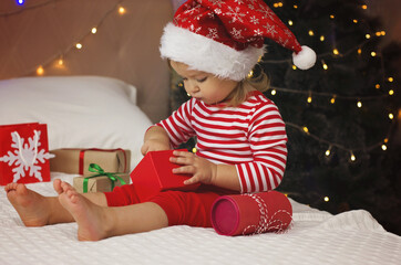 Wall Mural - Happy Holidays! Adorable baby girl under Christmas tree with gift boxes. Christmas present. Cure girl in Santa hat open gift box.