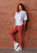 © oleg_ermak - Fashion style studio portrait of beautiful red-haired young girl in leggings pants and white t-shirt. Model standing and posing beside brick wall