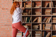 © oleg_ermak - Redhead female college student taking book from shelf in library. Stands on the stairs to get a book from the top. Side view, copy space.