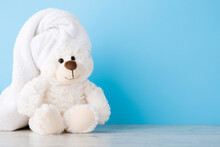Smiling, Lovely White Teddy Bear Sitting On Shelf At Light Blue Wall. Towel On Head. Closeup. Children Bathing Concept. Pastel Color. Empty Place For Text. Front View.