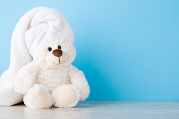 Smiling, lovely white teddy bear sitting on shelf at light blue wall. Towel on head. Closeup. Children bathing concept. Pastel color. Empty place for text. Front view.